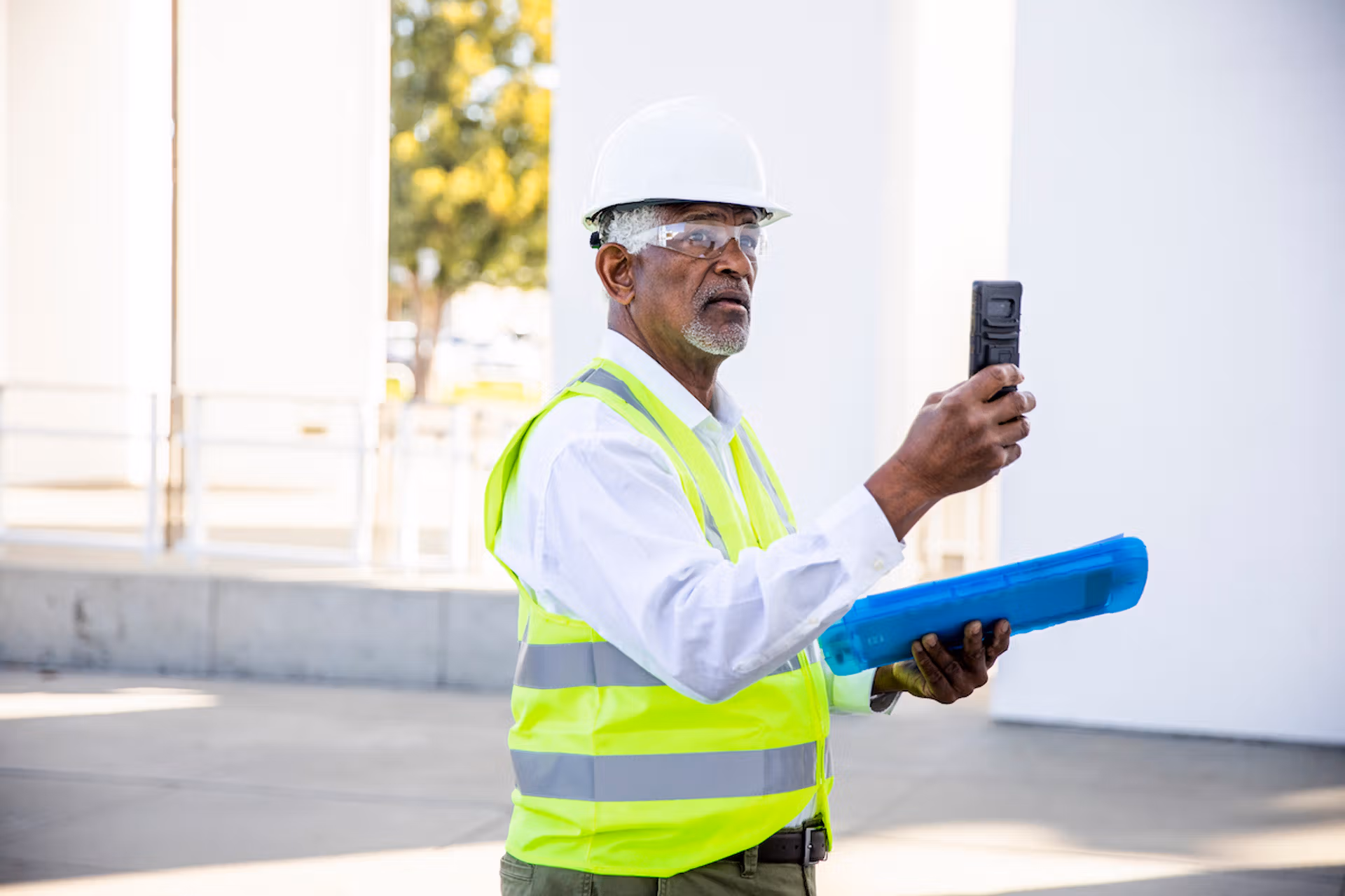 An older man wearing a hard hat and safety vest, holding a clipboard, stands in a construction site environment.