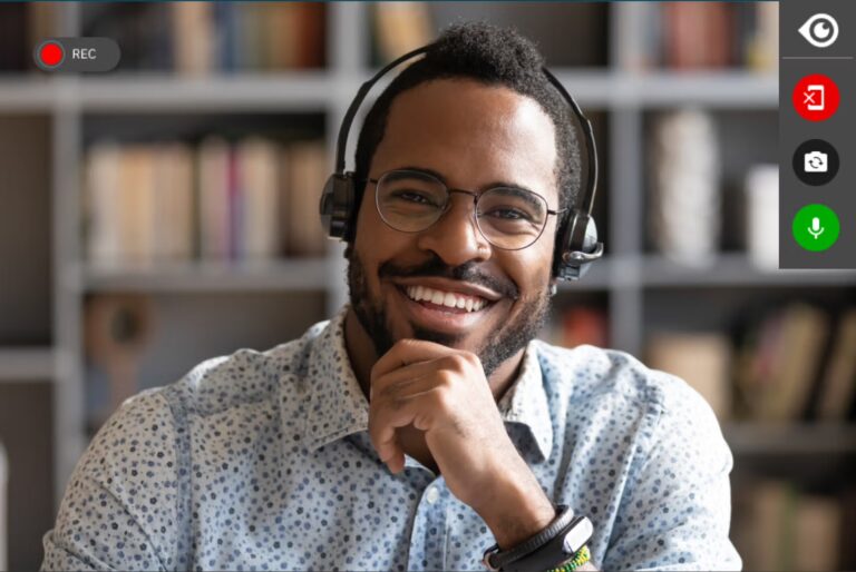 A smiling man wearing headphones sits in front of a computer screen, engaged and enjoying his activity.