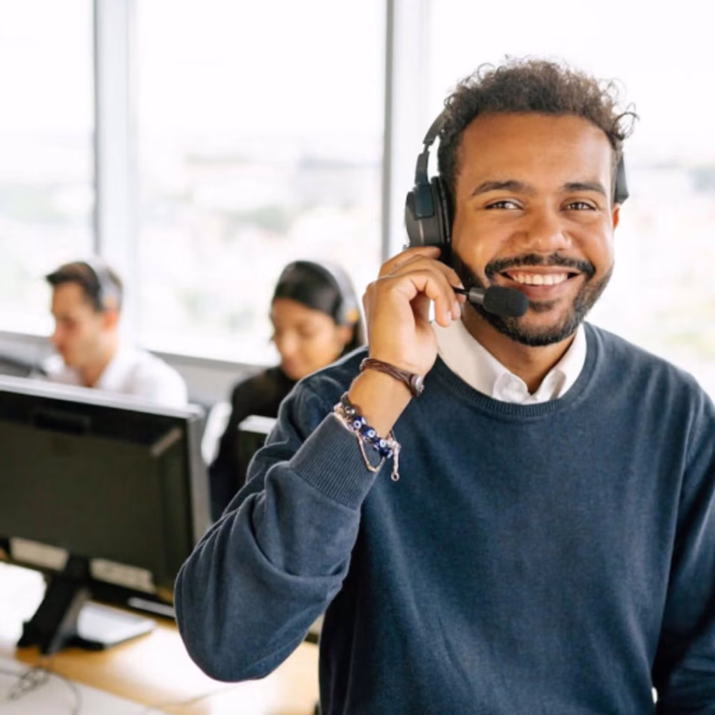 A smiling man wearing a headset, seated at a desk in a modern office environment.
