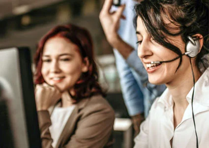 Two women wearing headsets collaborate in a modern office environment, focused on their tasks.