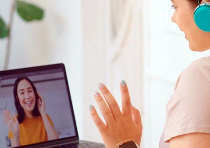 A woman wearing headphones sits in front of a laptop displaying another woman on the screen, engaged in a virtual conversation.