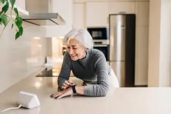 An older woman checks her smartwatch while preparing food in a well-lit kitchen, showcasing modern technology in daily life.
