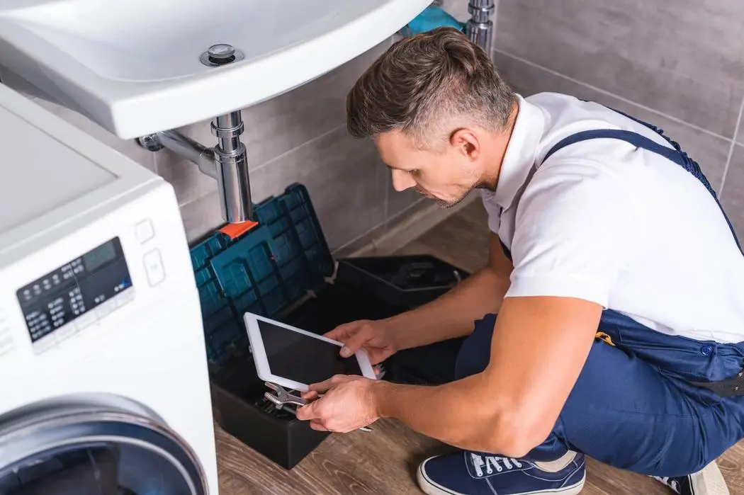 A man uses a tablet to assist him in fixing a washing machine in a home setting.