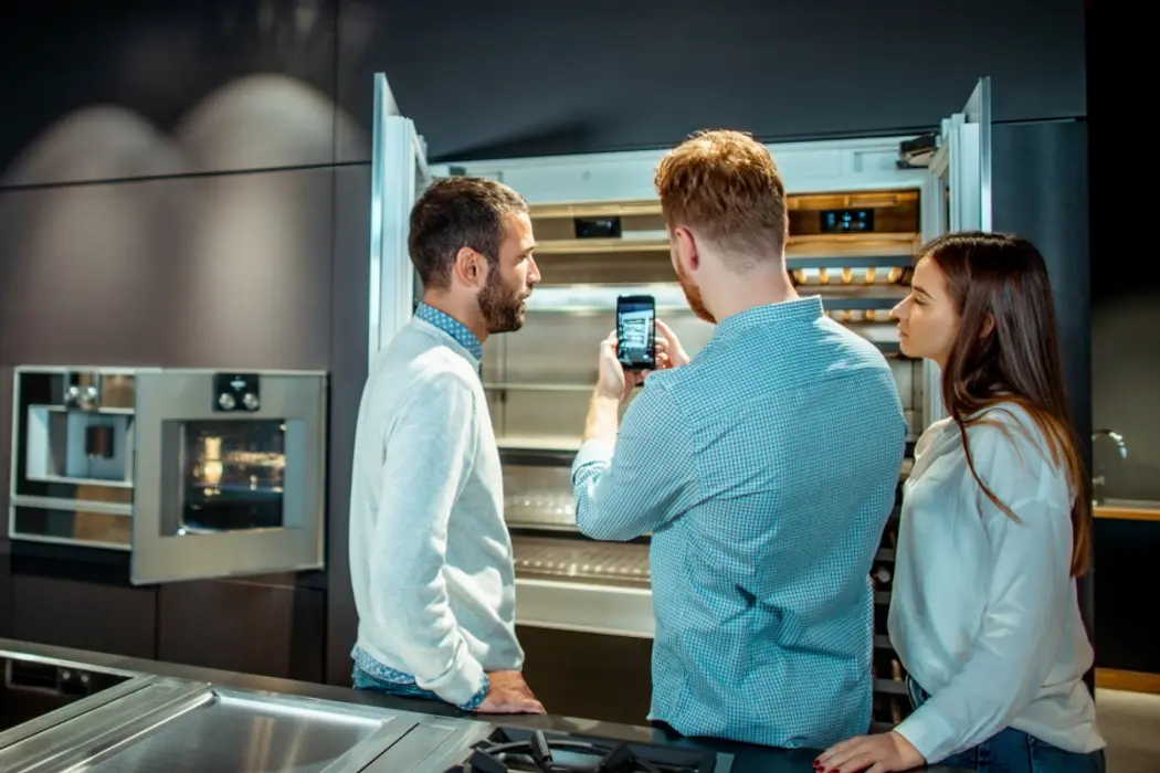 In front of a refrigerator, three individuals are engaged in looking at a cell phone, sharing a moment of interest.