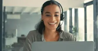 A woman with a headset smiles while engaging with her laptop, showcasing a positive and focused work environment.