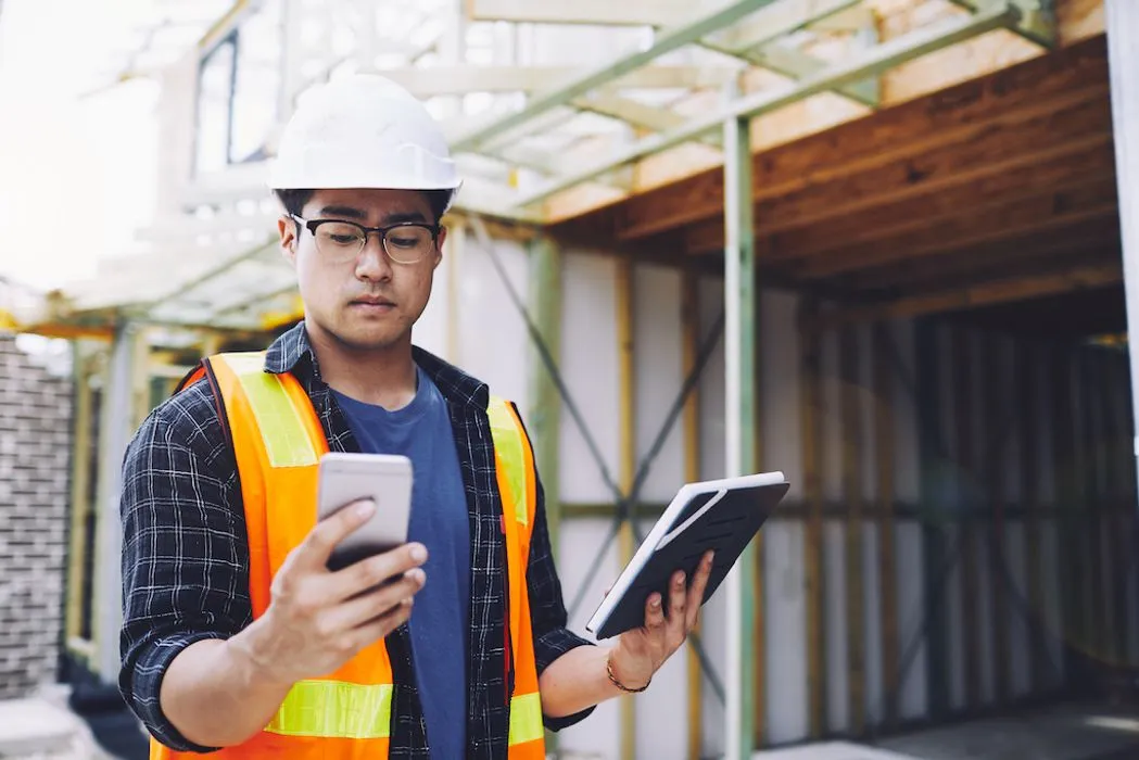 A man wearing a hard hat and safety vest is holding a tablet, focused on his work in a construction environment.
